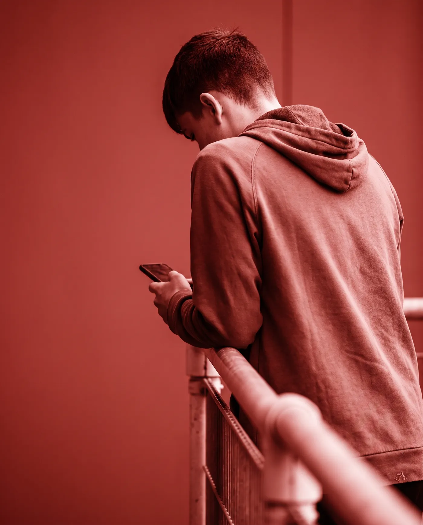 Ein Fußballer lehnt am Geländer des Platzes und schaut auf sein Handy, von hinten